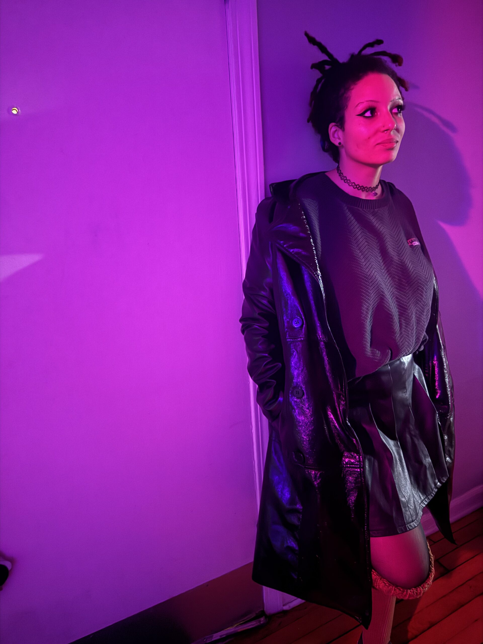 Picture of site creator, black women in purple lighting standing against a wall wearing a trench coat.