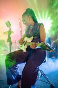 Black Women with purple jumpsuit playing guitar on stage with stage lights and mic.